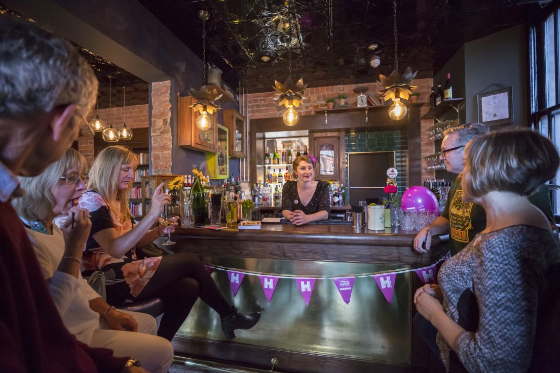 An image of people sitting in a circle in front of a bar in a pub, holding drinks and talking to the woman standing behind the bar.