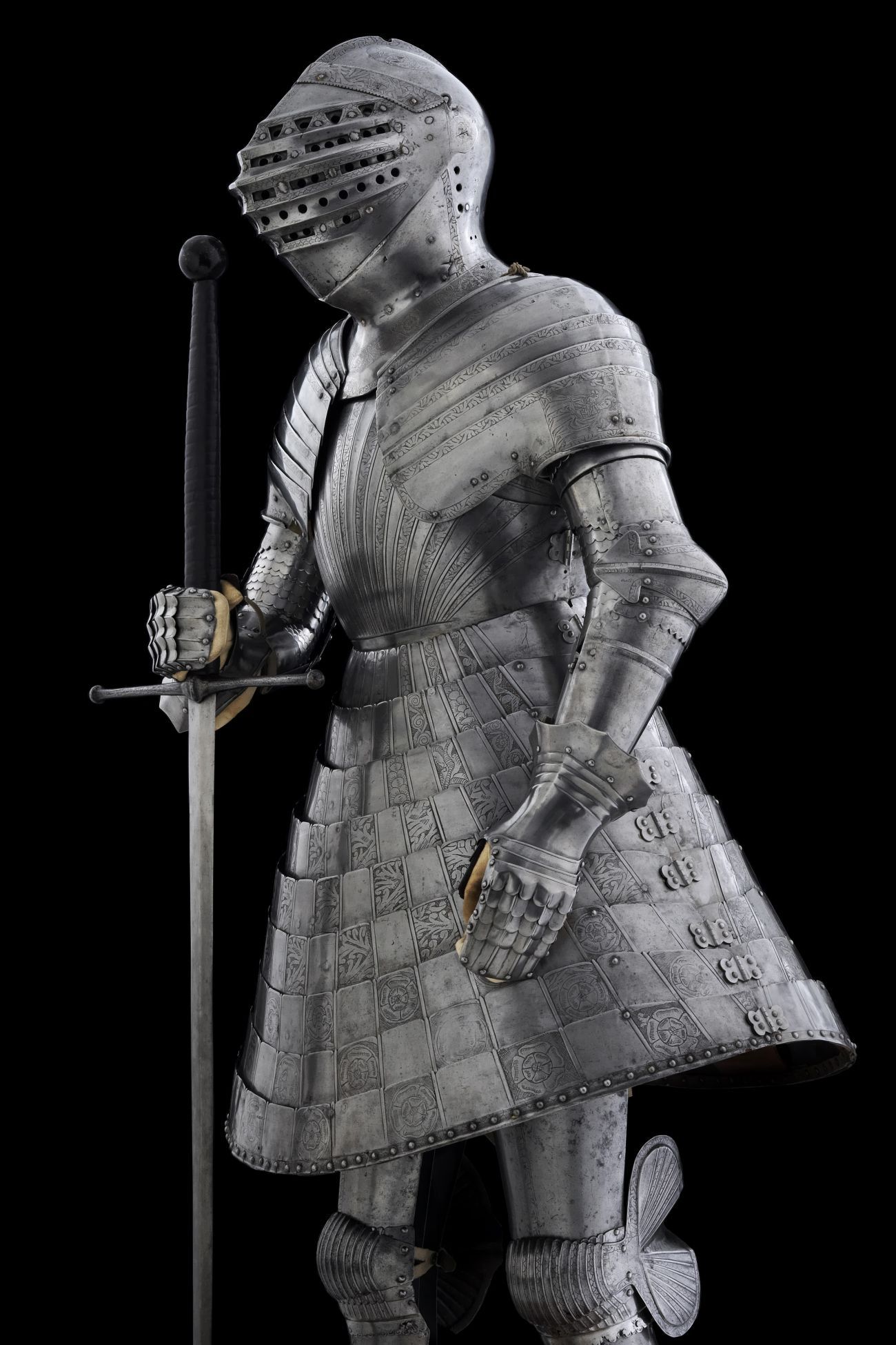 Full suit of armour with helmet and hooped metal skirt. Stood upright with gauntleted hand grasping the hilt of a huge sword pointing to the floor. 