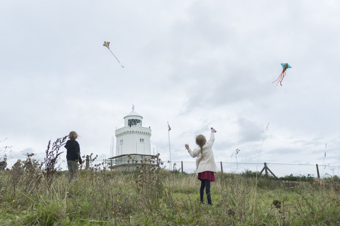 Two children playing in a grassy field with a lighthouse, both flying kites.